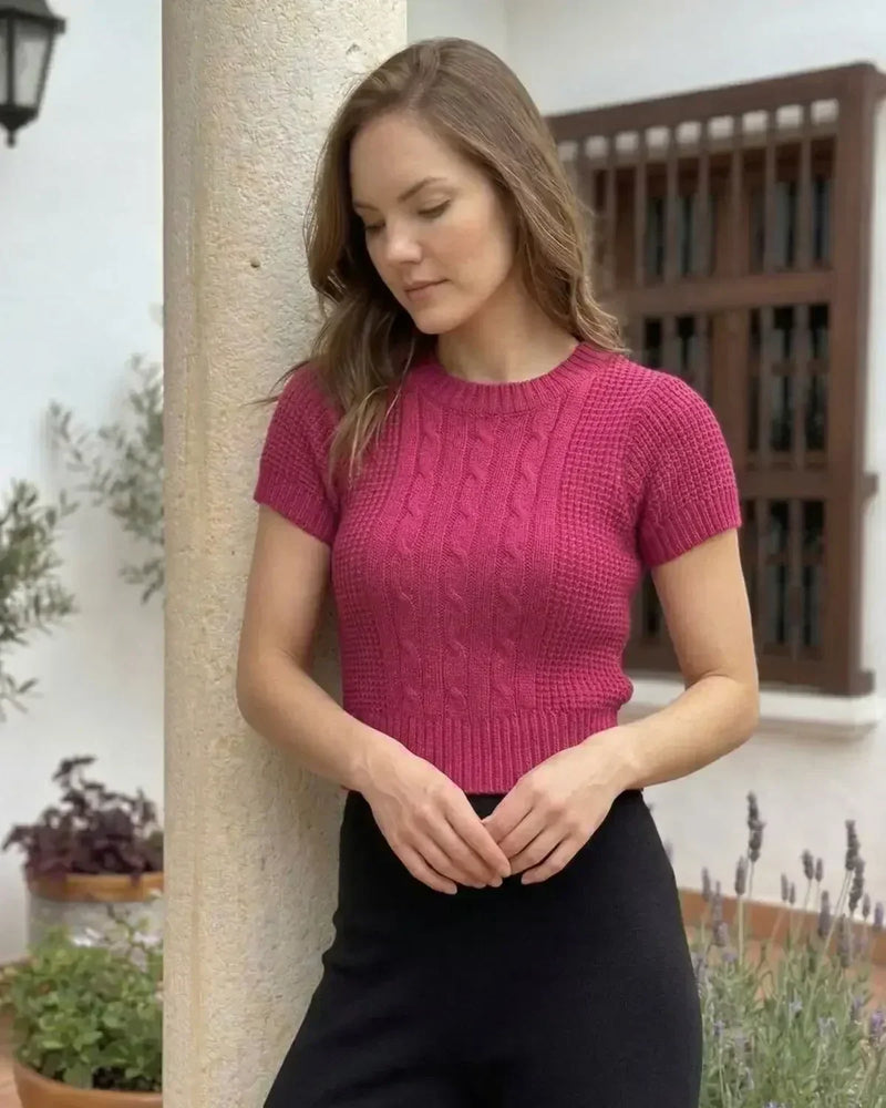Mujer con suéter tejido rosa de manga corta y pantalón negro, en jardín elegante.