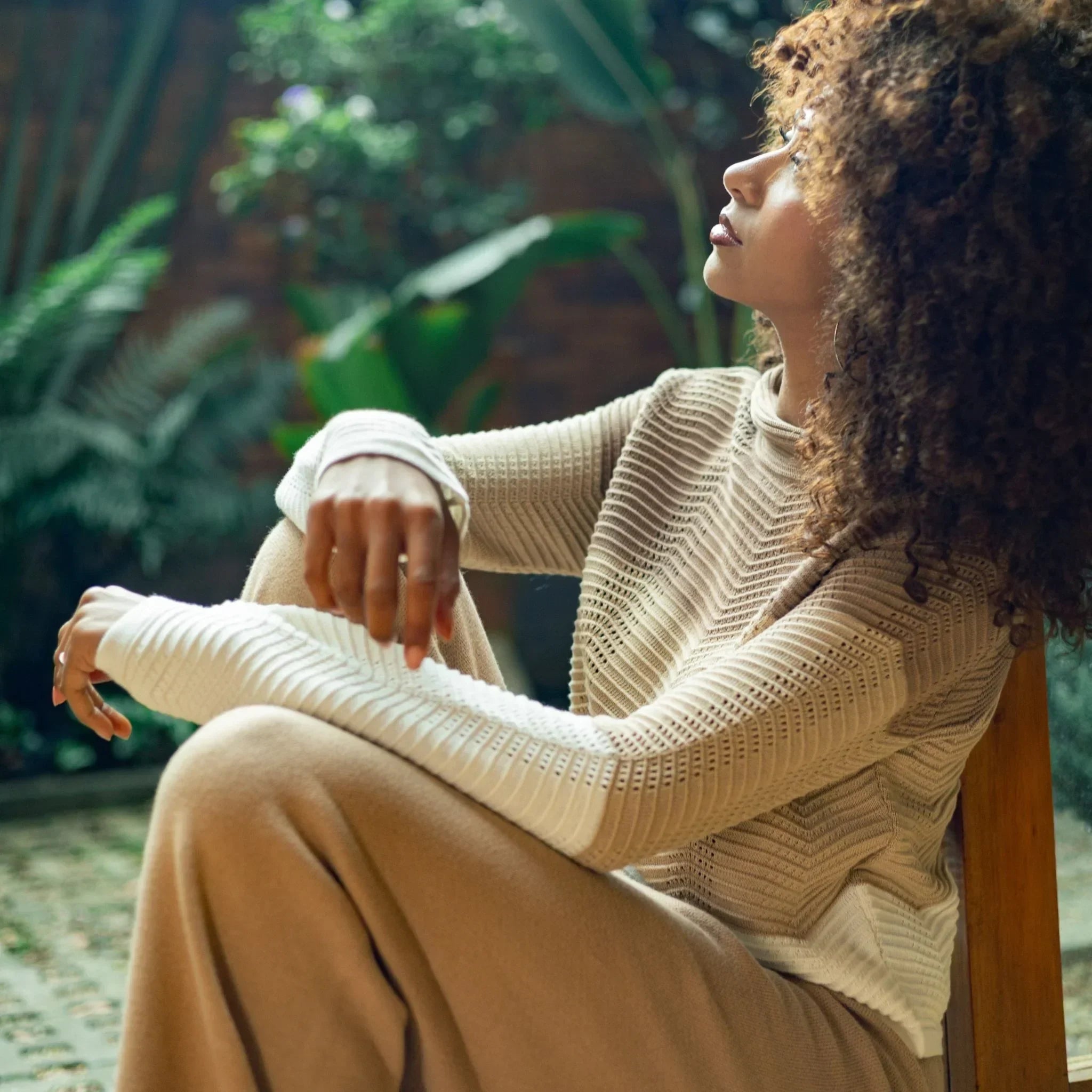 Mujer con cabello rizado usando suéter tejido beige y pantalón amplio, sentada en un jardín