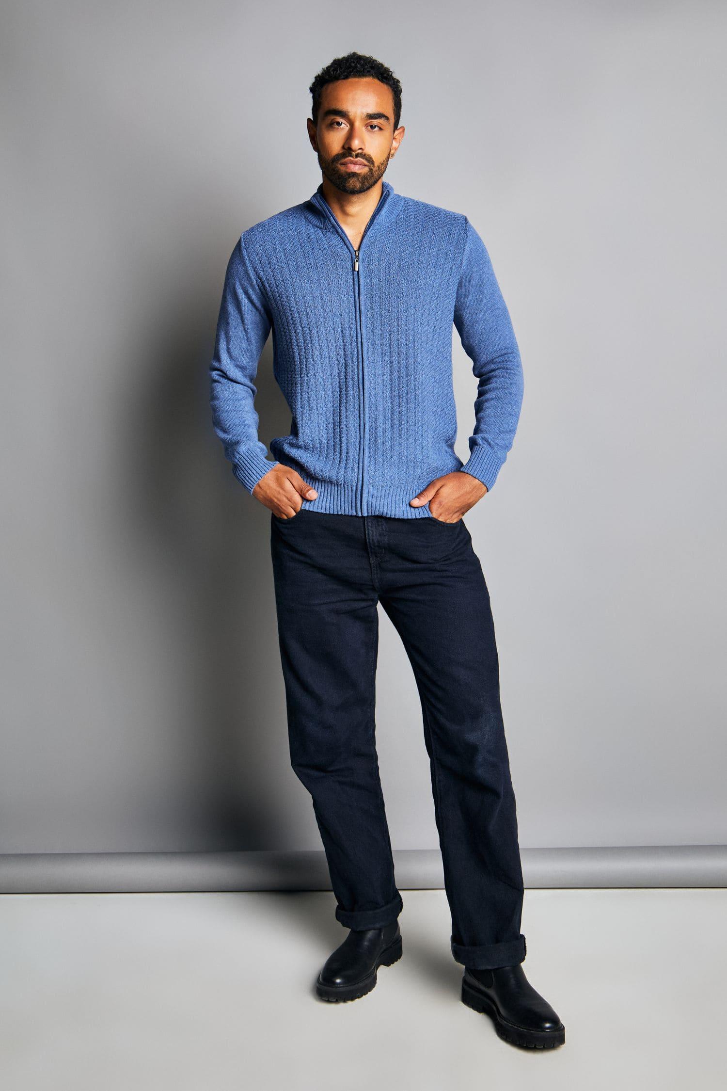 Hombre con chaqueta tejida azul, pantalón oscuro y botas negras, fondo gris.