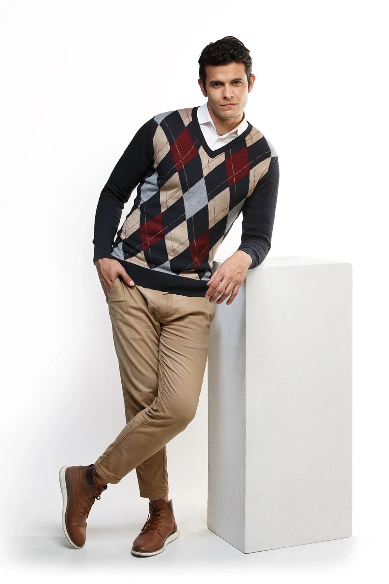 Hombre con suéter de rombos tejido, pantalón beige y botas café posando en estudio