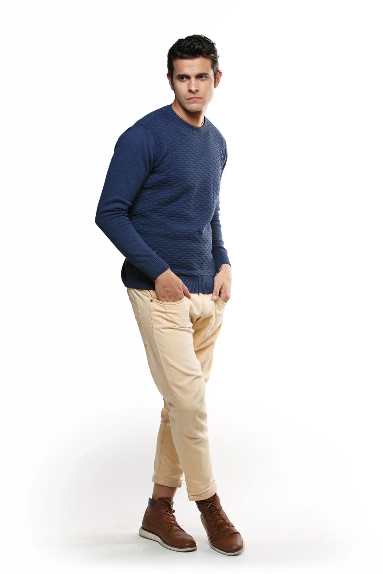 Hombre con suéter tejido azul marino, pantalón beige y botines marrones, fondo blanco.
