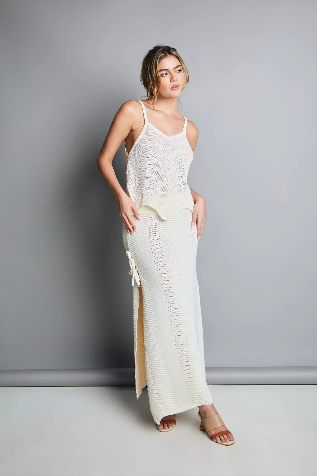 Mujer con conjunto tejido blanco, falda larga con abertura lateral y top de tirantes, fondo gris