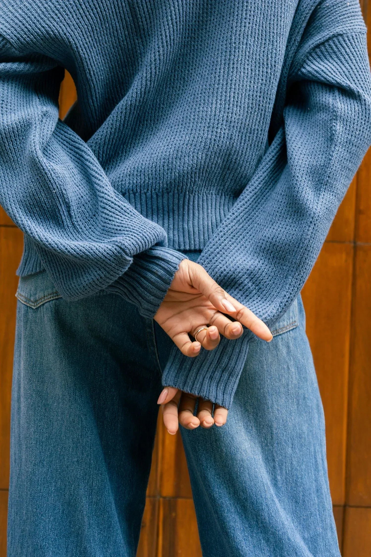 Mujer con suéter tejido azul y pantalón de mezclilla, de espaldas, fondo madera.