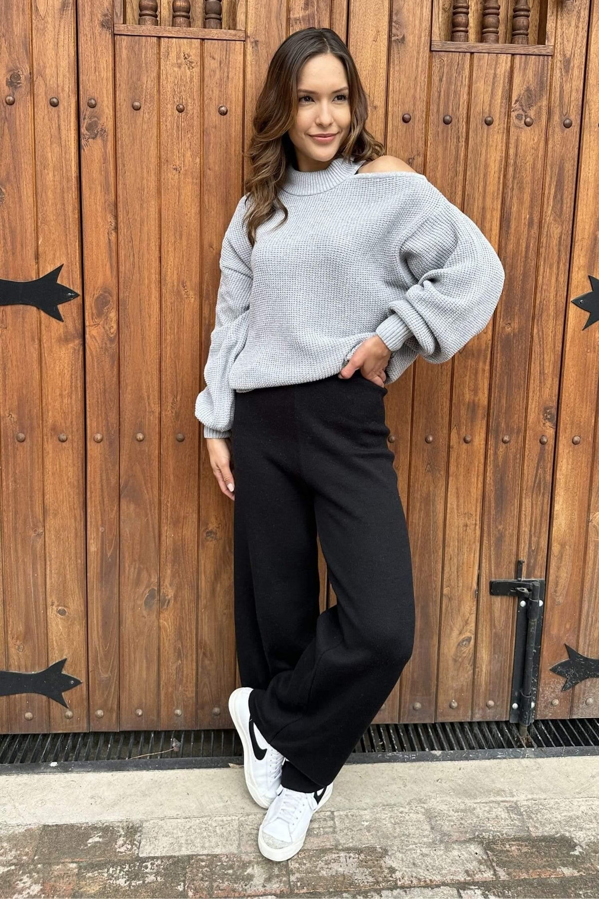 Mujer joven con suéter gris de hombro descubierto y pantalón negro, estilo casual, frente a una puerta de madera.