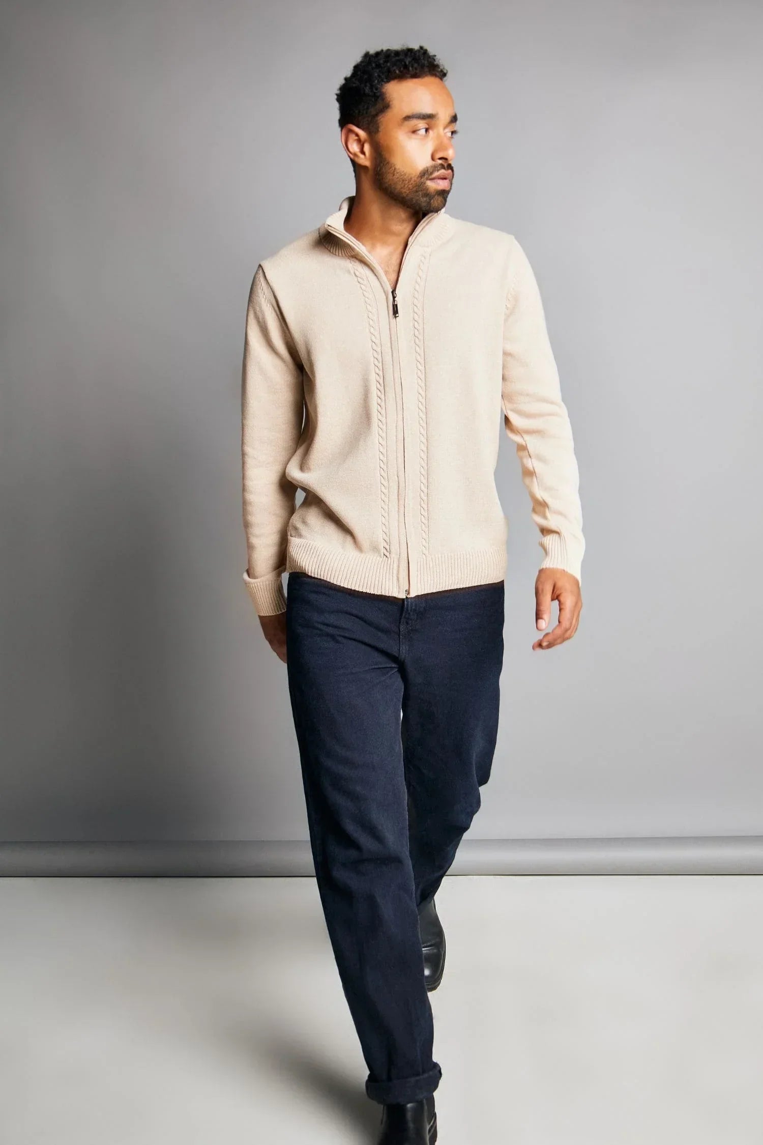 Hombre con suéter beige tejido de punto y pantalón azul oscuro, fondo gris, moda masculina.
