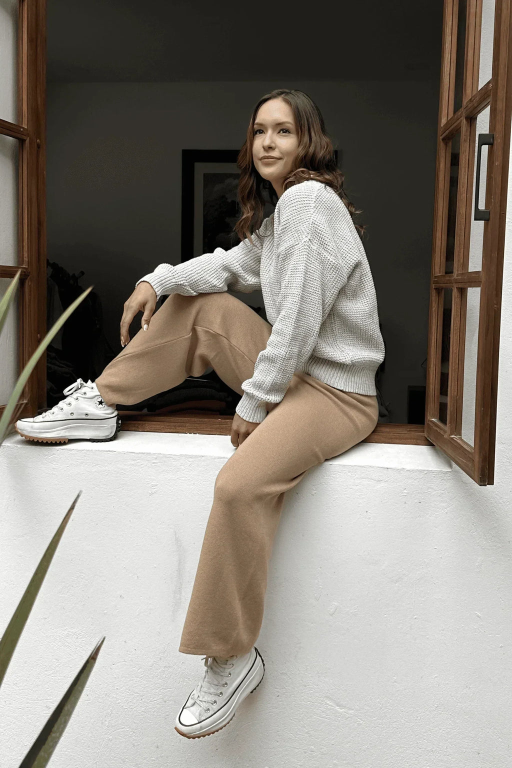 Mujer con suéter gris y pantalón beige sentada en una ventana, looks cómodos y casuales