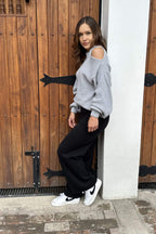 Mujer con suéter gris de hombro descubierto, pantalón negro y tenis blancos estilo casual.