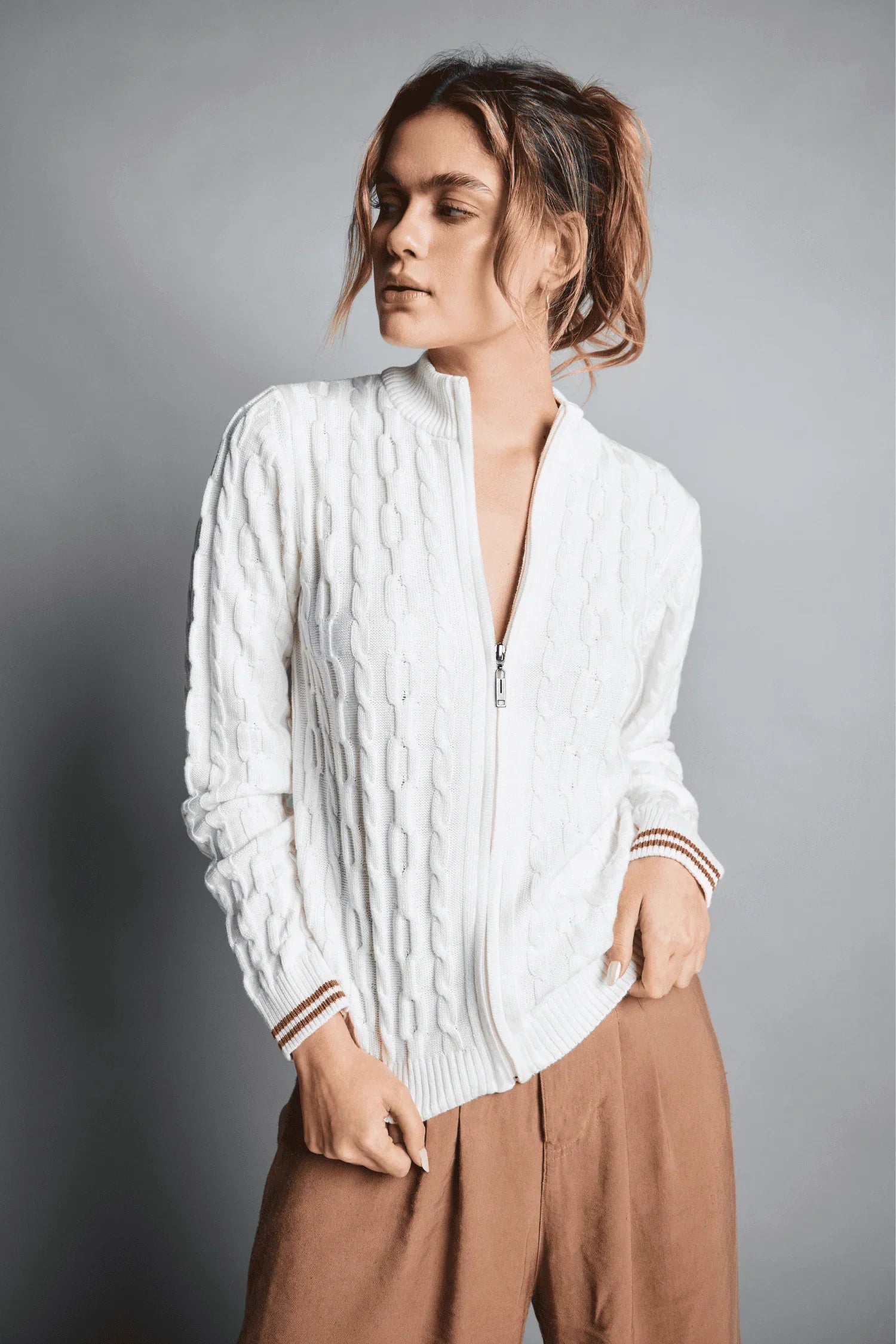 Mujer con chaqueta tejida blanca con cremallera y pantalón marrón, fondo gris