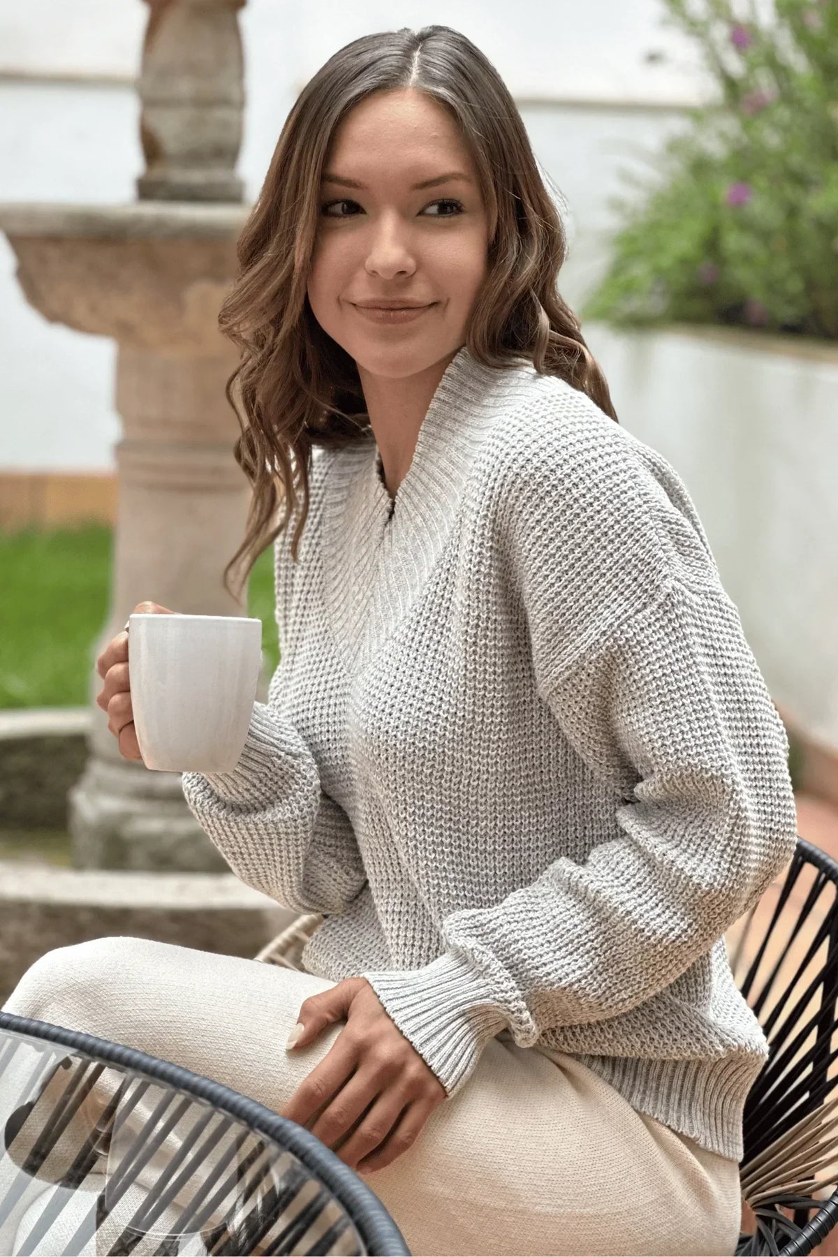 Mujer sentada con suéter tejido gris claro, sosteniendo una taza en un ambiente al aire libre.