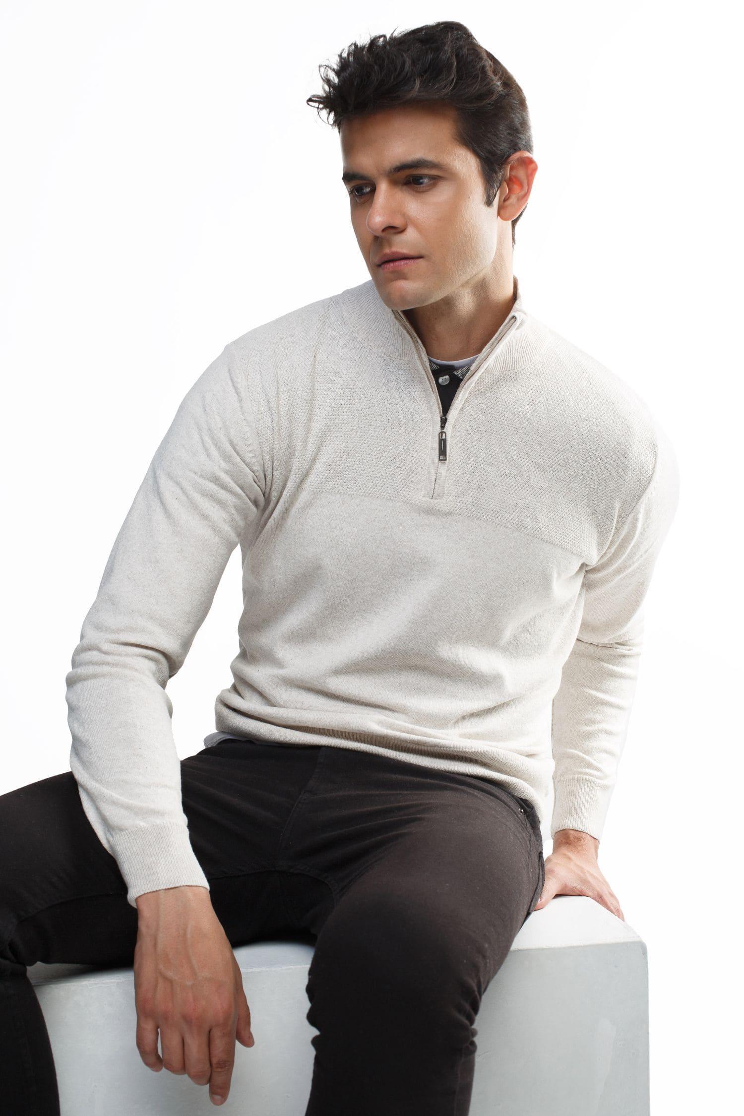 Hombre joven con suéter de punto beige y cremallera corta, sentado sobre fondo blanco.