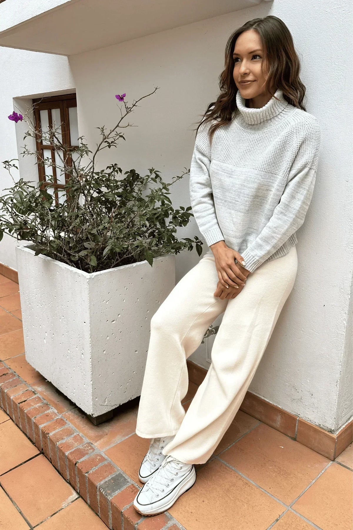Mujer con suéter gris claro de cuello alto, pantalón crema y zapatillas blancas, estilo casual