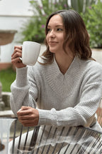 Mujer sentada al aire libre con suéter gris tejido, sosteniendo una taza blanca.
