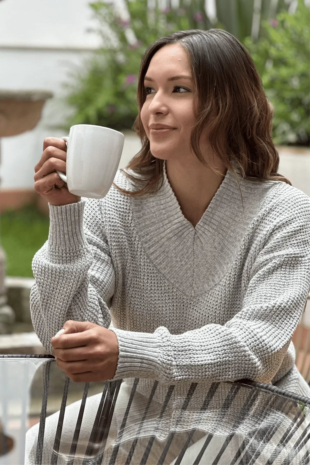 Mujer sentada al aire libre con suéter gris tejido, sosteniendo una taza blanca.