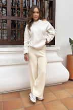 Mujer con suéter y pantalón beige holgados, look casual, fondo exterior de casa blanca