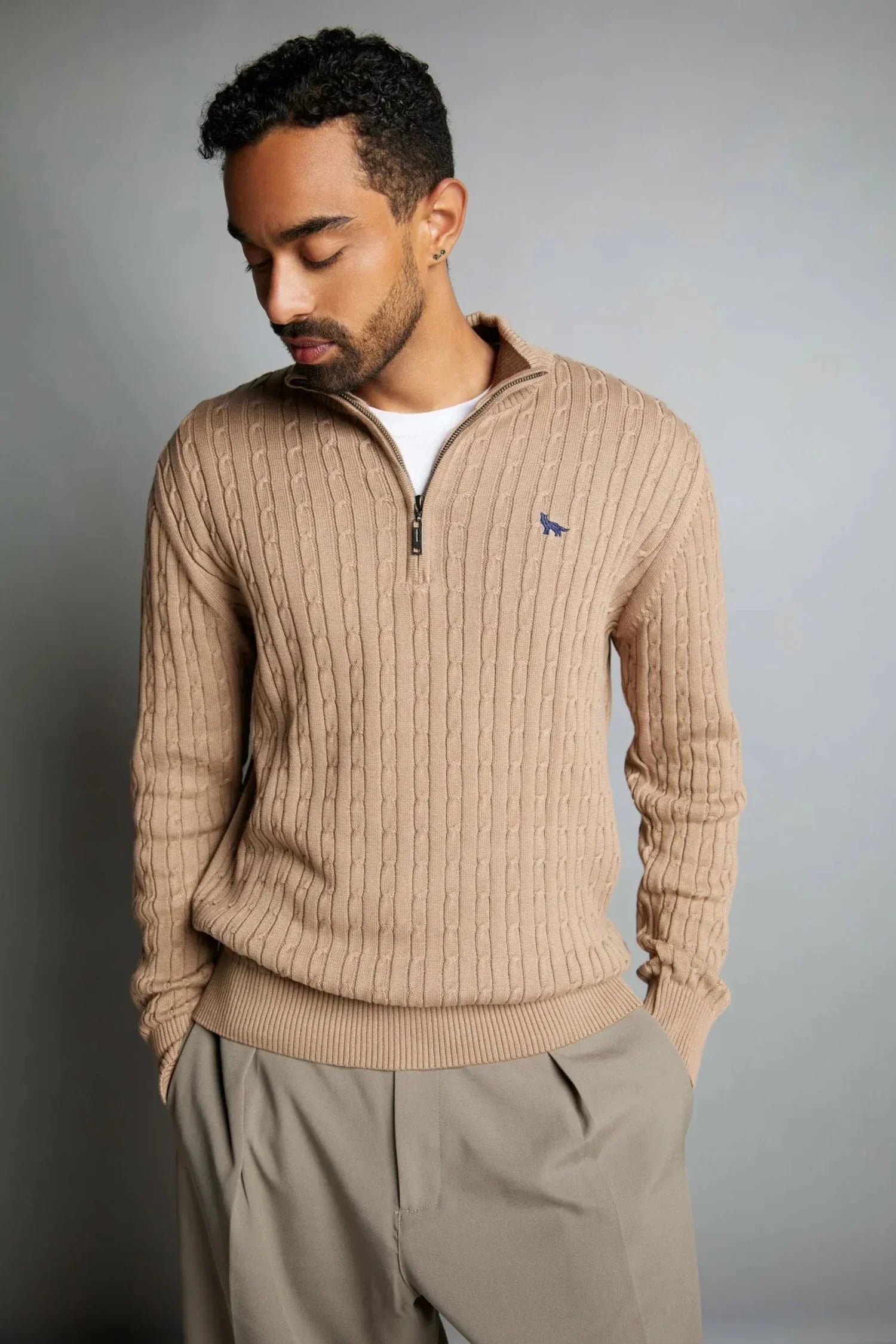 Hombre con buzo tejido beige de punto, cierre corto, detalle bordado azul, moda casual.