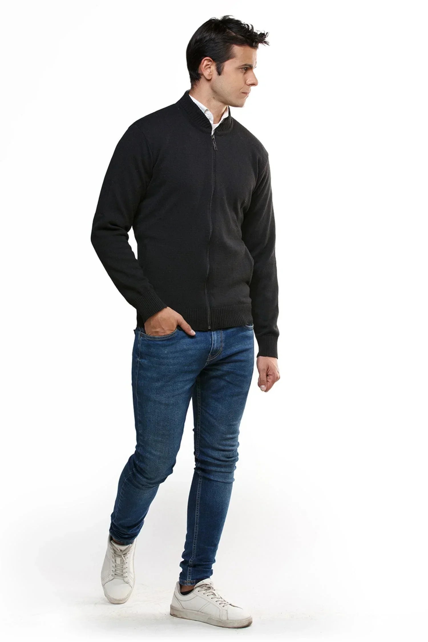 Hombre con chaqueta negra de punto, jeans azul y tenis blancos sobre fondo blanco