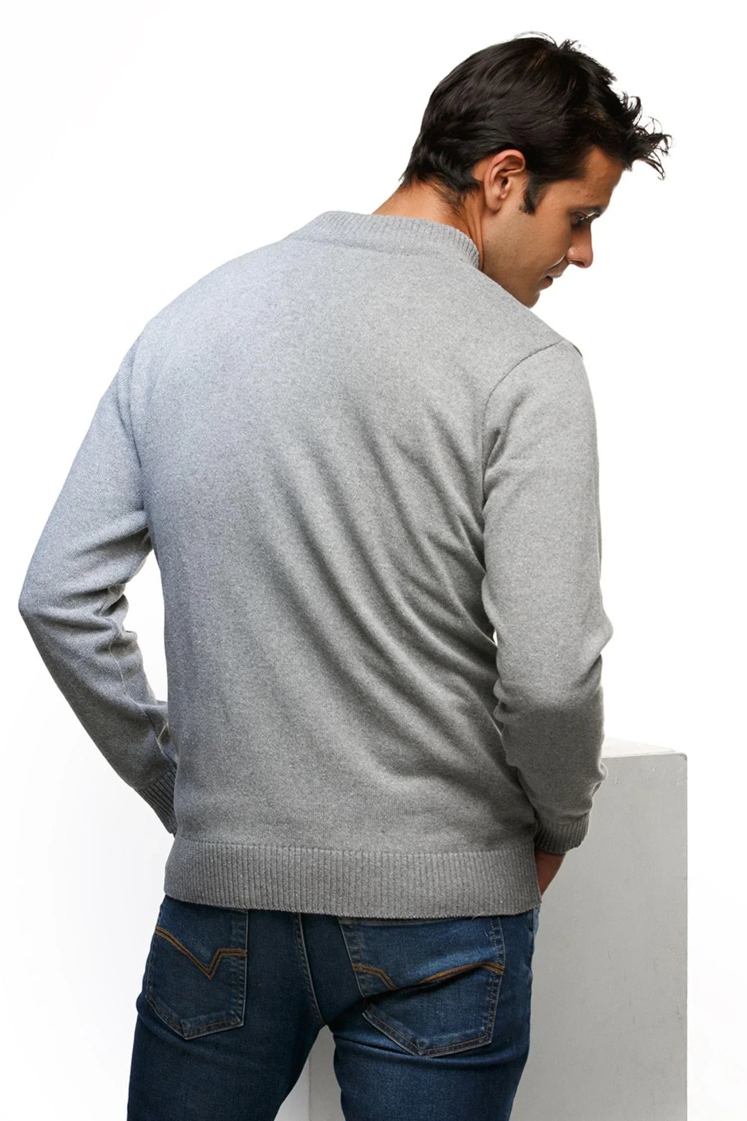 Hombre de espaldas con suéter gris de punto y jeans azul, fondo blanco, moda masculina.