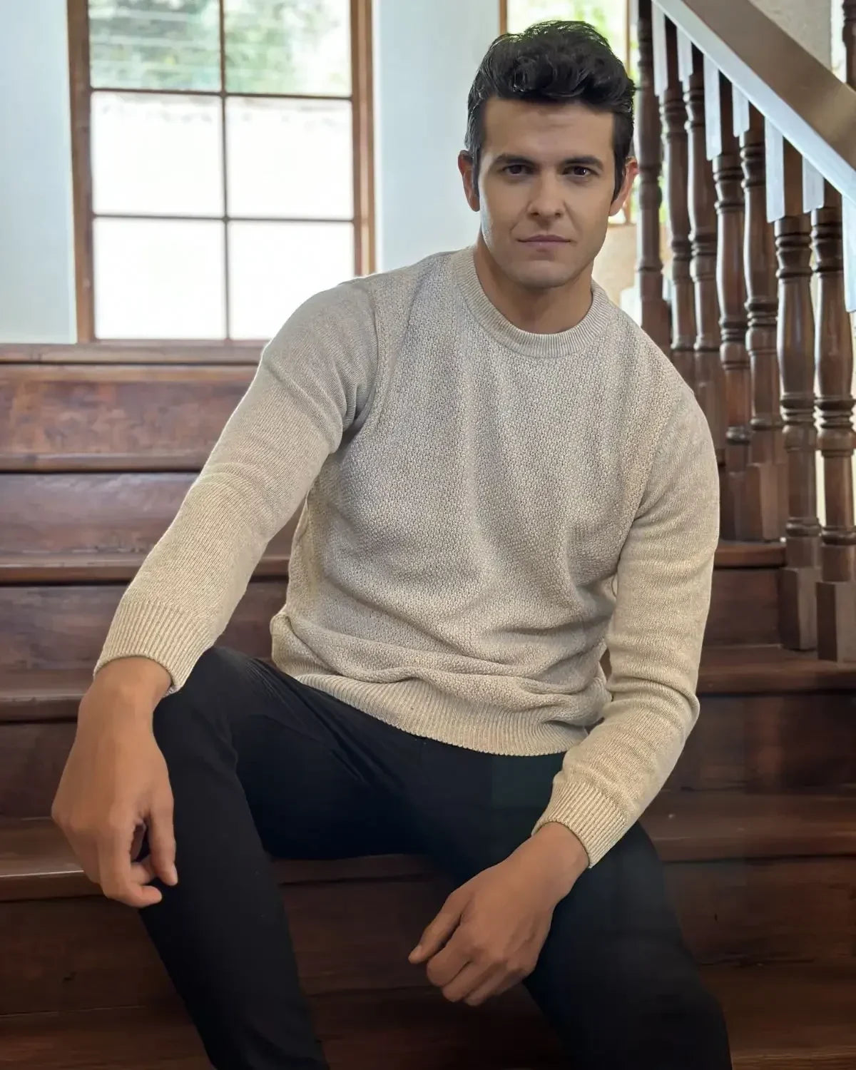 Hombre sentado en escalera con suéter beige y pantalón negro, moda casual masculina.