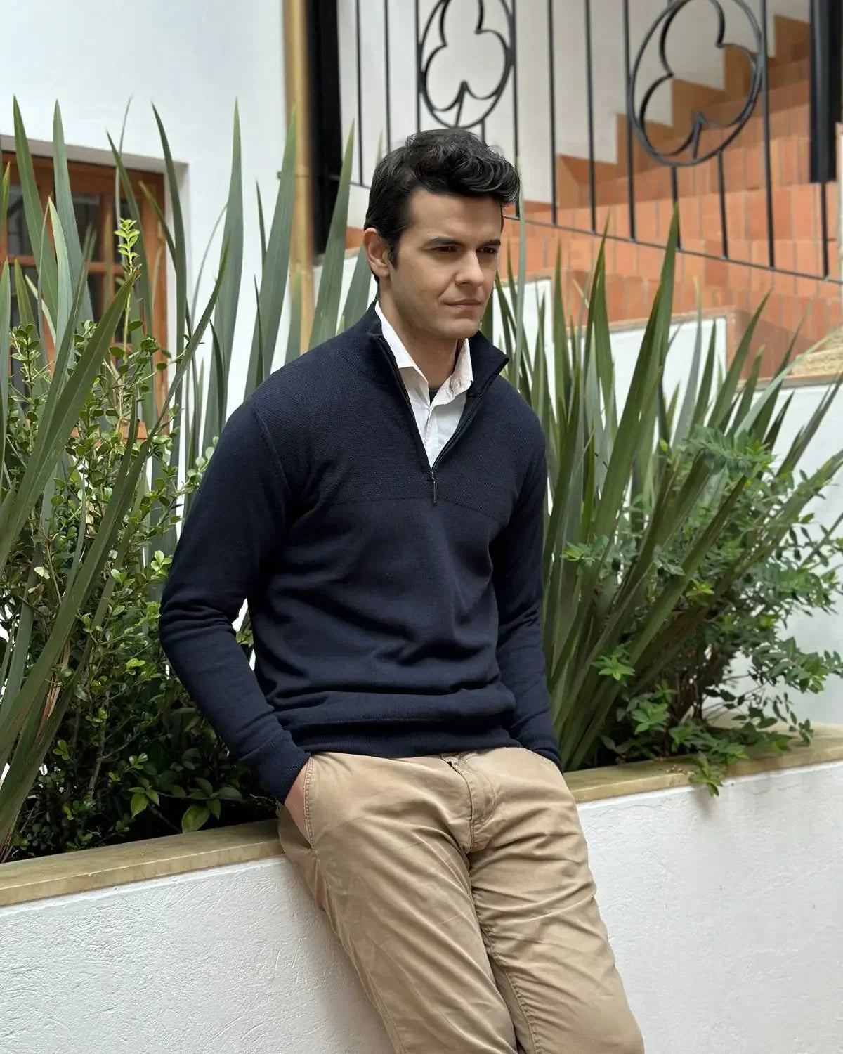 Hombre con suéter azul marino y pantalón beige posando en jardín con plantas y fondo urbano