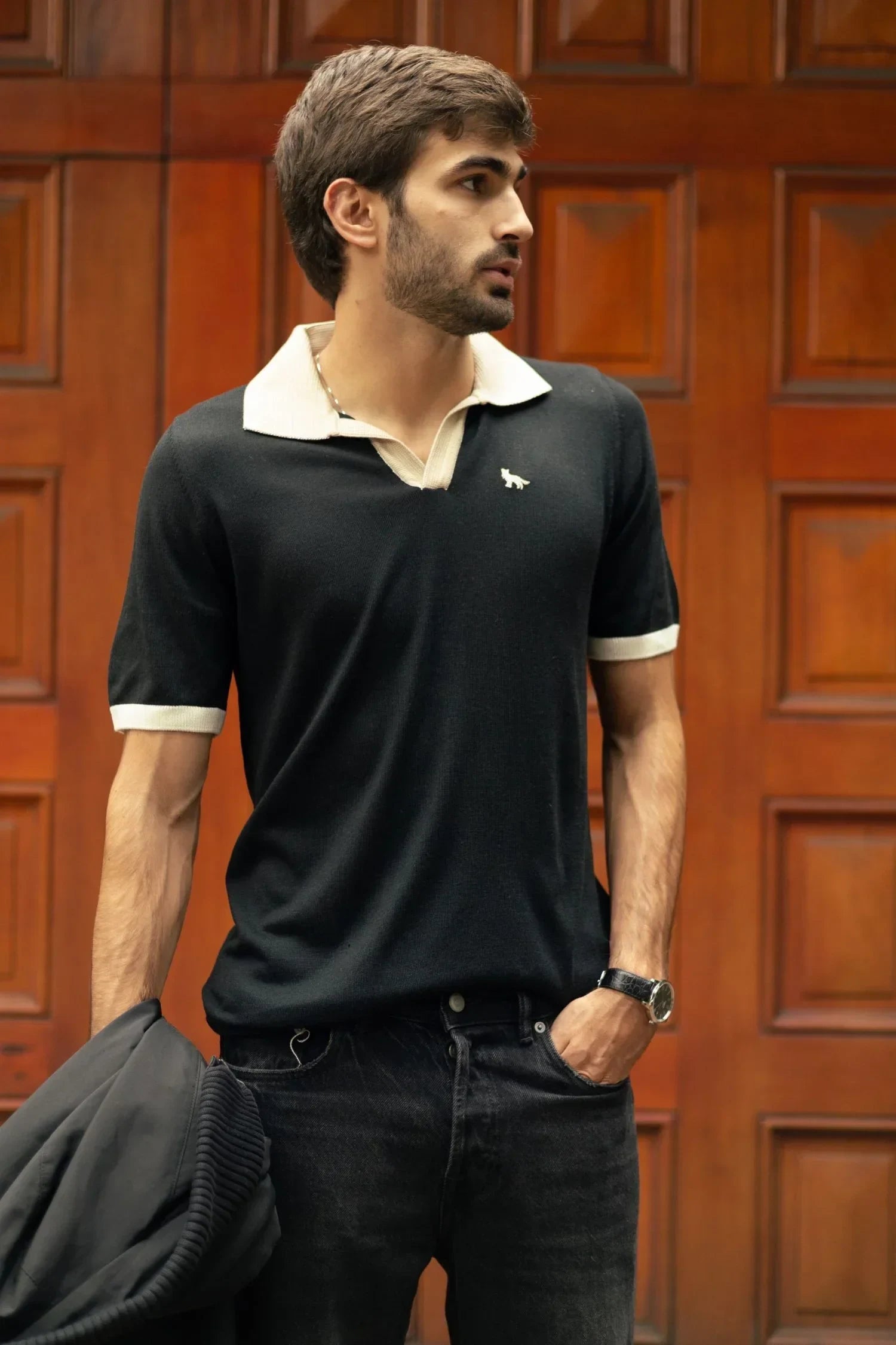 Hombre con polo negro de manga corta, cuello beige y pantalón oscuro frente a puerta de madera.